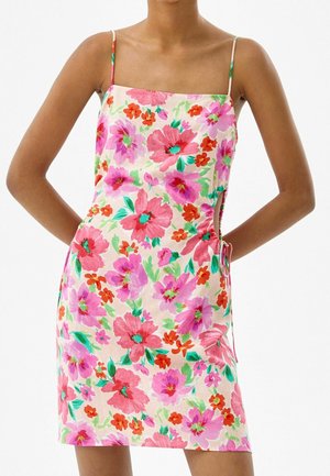 Hihaton mekko, jossa ohuet olkaimet ja vaalealla taustalla vaaleanpunainen, punainen ja violetti kukkakuosi, pukeutuneena henkilö, jonka kädet ovat selän takana.
