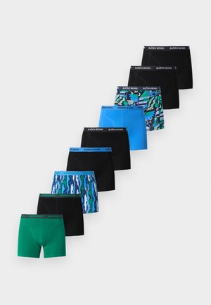 Großer, schlanker Mann, barfuß auf einem Teppich stehend, trägt schwarze Boxershorts mit grünem Bund, blickt nach links vor einem schlichten hellen Hintergrund.