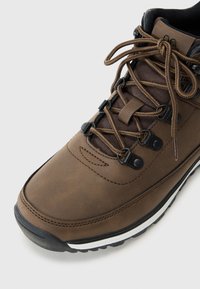 Scarpa da trekking marrone con tomaia in sintetico liscio, lacci scuri, occhielli neri e accenti bianchi sulla suola. Design robusto e testurizzato.
