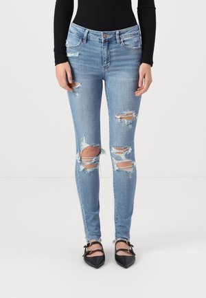 Person wearing light blue ripped skinny jeans, black pointed-toe shoes with straps, and a black long-sleeve top, standing on white floor.