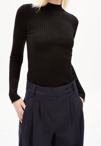 Femme portant un haut noir côtelé à manches longues rentré dans un pantalon plissé taille haute bleu marine, les mains dans les poches, sur un fond blanc.