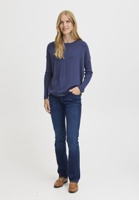 Pulover lung de culoare albastru marin, cu un croi lejer, guler rotund și detalii subtile de cusătură, asortat cu blugi de culoare albastră închisă și pantofi maro.