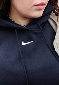Námornícky modrá mikina s bielym logom Nike. Tkanina vyzerá jemne, s šnúrkou a textúrovaným povrchom. Sveter svetlej farby je čiastočne viditeľný.