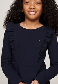 Abito a coste blu navy con maniche lunghe, dotato di dettagli arricciati lungo il petto e un piccolo logo sul lato sinistro.