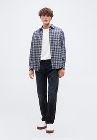 Νέος άνδρας με σγουρά μαλλιά, φορώντας μπλε πουκάμισο με καρό κουμπιά πάνω από λευκό μπλουζάκι, σκούρα τζιν και λευκά αθλητικά παπούτσια, στέκεται μπροστά σε απλό φόντο.