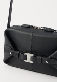 Bolsa de couro preta com uma forma estruturada, apresentando um fecho de zíper, detalhe decorativo com alça e fivela, textura suave e costura em contraste.