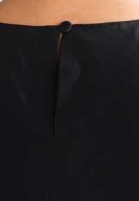 Černé šaty z pohledu zezadu s vertikálním rozparkem a kulatým černým knoflíkem na zapínání u krku, vyrobené z hladkého, matného materiálu.