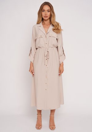 Kobieta stojąca twarzą do przodu, ubrana w beżową midi sukienkę na guziki z długimi rękawami, posiadającą kieszenie i pasek w talii, oraz beżowe sandały na obcasie z paskami.
