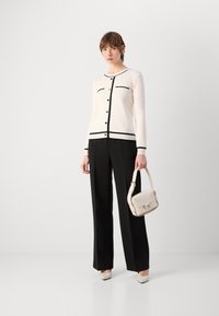 Gilet blanc avec des bordures et des boutons noirs, associé à un pantalon large noir. Tenant un petit sac à main blanc et portant des talons aiguilles.