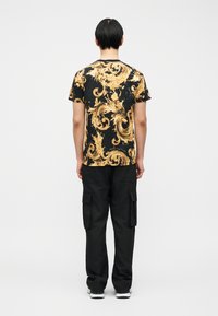 Άτο μο με κοντά μαύρα μαλλιά στέκεται με την πλάτη γυρισμένη, φορώντας μια μαύρη μπλούζα με χρυσά περίτεχνα σχέδια και μαύρο παντελόνι cargo με αθλητικά παπούτσια.