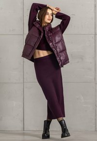 Gepolsterte Weste in Dunkelviolett mit hohem Kragen, vorderen Taschen und einer glänzenden Textur, getragen über einem passenden, figurbetonten Oberteil und Rock.