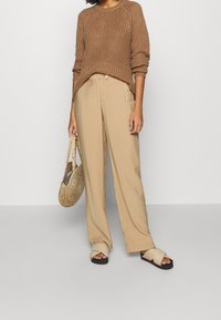 Brauner Strickpullover, beige weit geschnittene Hose, schwarze Riemchensandalen und eine geflochtene Tasche. Texturierte Materialien und lockere Passform.