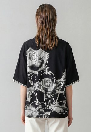 Persoon met nat, halflang haar die een zwart shirt draagt met een groot wit pixelachtig rozenontwerp op de rug en witte broek.