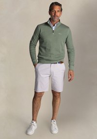 Groene pullover met een ritskraag, lichtgrijze shorts met een gemêleerd patroon, en witte sneakers. Het geheel heeft een blauw-wit gestreepte riem.