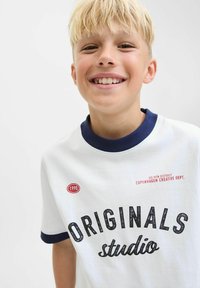 Bijela pamučna majica s mornarsko plavim ovratnikom, crnim natpisom "ORIGINALS studio" i crvenom oznakom "1990". Dizajnirana za svakodnevno nošenje.