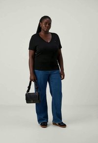 Woman stands wearing a black V-neck shirt, blue jeans, black shoes, and holds a black handbag against a plain light background.