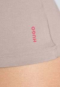Beige stretchshorts med en myk tekstur, med en synlig rød "HUGO"-logo på siden og pent syede sømmer langs kanten.
