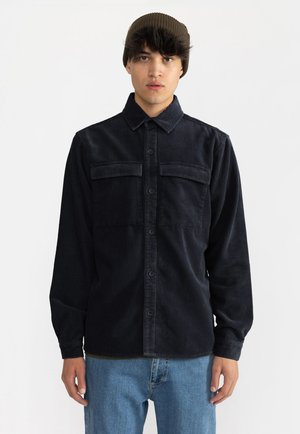 Zwarte ribfluwelen overhemden met een knoopsluiting, twee borstzakken en lange mouwen, gedragen over lichtblauwe denimjeans.