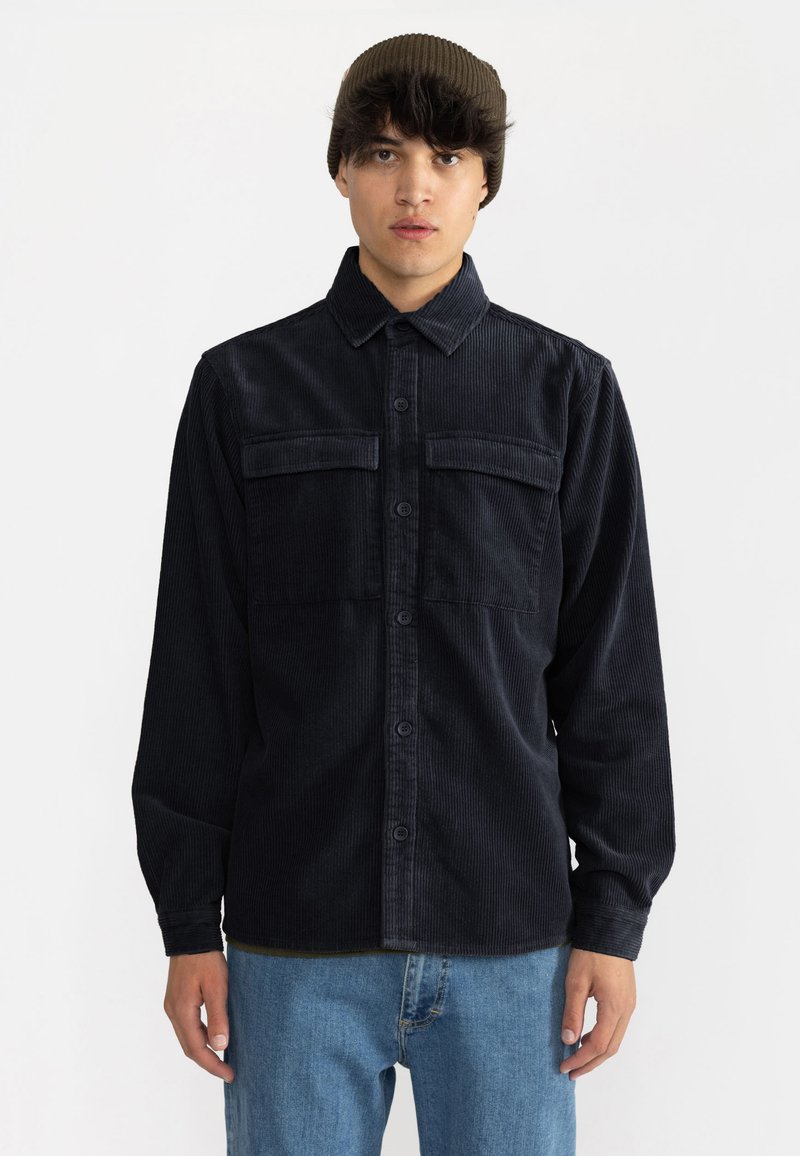 Sort kordfløjlsskjorte med knaplukning, to brystlommer og lange ærmer, båret over lyseblå denimjeans.