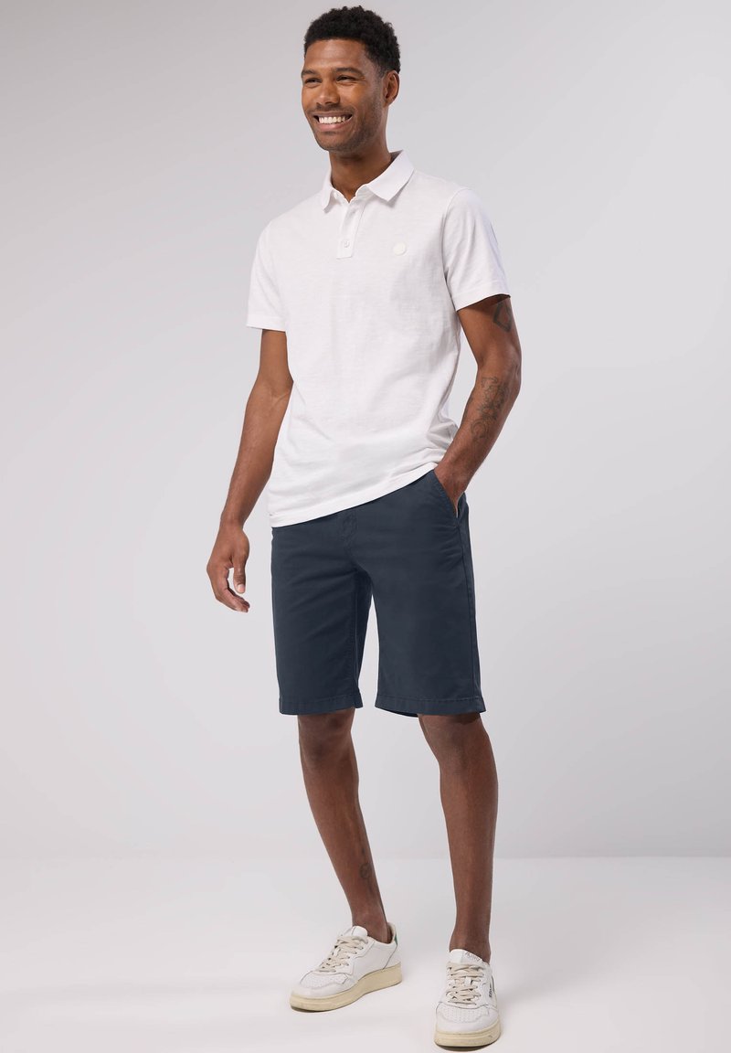 Vit bomullspolo med krage, kombinerad med marinblå shorts och vita sneakers. Klädseln har en avslappnad passform och minimala designelement.