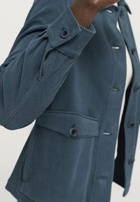 Modrá kordurová bunda s knoflíkovou přední částí, dvěma kapsami a ohrnutými rukávy s knoflíkovým detailem. Tkanina má jemně řasenou strukturu.