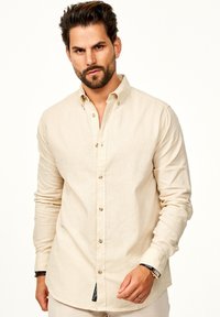 Hellbeige Leinenhemd mit langen Ärmeln, ausgestattet mit einem spitzen Kragen, Knopfverschluss und subtiler Textur. Etikett am Saum sichtbar.