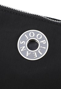 Černý nylonový pouzdro se stříbrným zipem a kulatým kovovým detaily s textem "JOOP! JEANS". Texturovaný povrch s viditelným šitím.