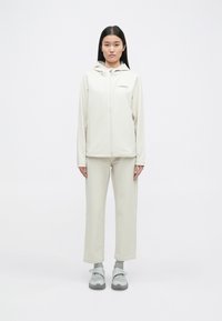 Veste zippée beige clair avec capuche, pantalon assorti et baskets grises. Présente un logo discret sur la poitrine et une texture lisse.