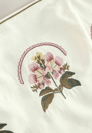 Weißer Stoff mit floraler Stickerei in Rosa, Gelb und Grün, mit einer gezackten rosa Verzierung über den Blumen.