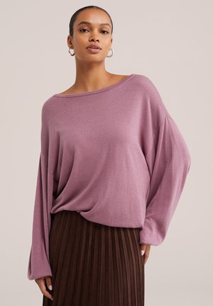 Femme portant un pull oversize mauve et une jupe plissée marron sur un fond beige clair.