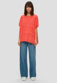 Koraal gestreept topje met korte mouwen, losse fit, gecombineerd met wijde blauwe jeans en witte plateau sneakers, op een neutrale achtergrond.