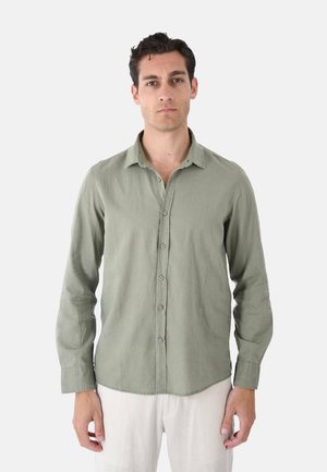 Uomo in piedi di fronte, indossa una camicia verde chiaro con bottoni e maniche lunghe e pantaloni bianchi su uno sfondo semplice.