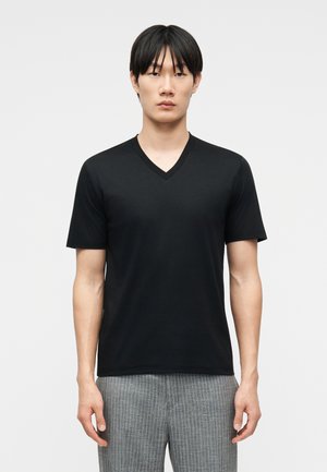 Νεαρός ενήλικας που φοράει μαύρο T-shirt με V λαιμόκοψη και κοντά μανίκια και γκρι παντελόνι με λεπτές ρίγες, στέκεται μπροστά σε απλό ανοιχτό γκρι φόντο.