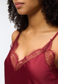 Γυναίκα που φοράει σκούρο κόκκινο σατέν καμίσω με δαντέλα και λεπτές τιράντες στους ώμους, εμφανίζοντας τον λαιμό και το πάνω μέρος του στήθους.