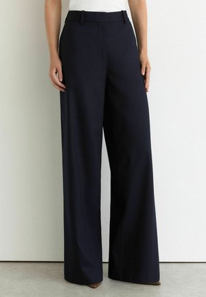 Femme portant un pantalon bleu marine taille haute à jambes larges avec passants de ceinture, debout devant un mur blanc uni et un sol gris.