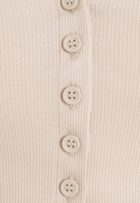 Cardigan côtelé en beige doux avec six boutons ronds à l'avant. Contrastes de texture notables entre le tissu et les boutons.