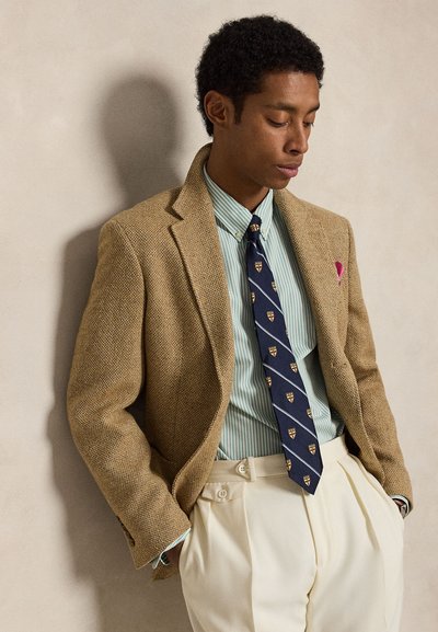 Homme portant un blazer en tweed beige, une chemise rayée vert clair, une cravate marine avec des motifs de blason, un pantalon plissé crème et un mouchoir rose, debout les mains dans les poches.