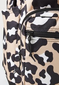 Sac à dos beige avec un imprimé léopard noir et blanc, poche zippée ouverte, tissu texturé et quincaillerie de zipper noire avec cordons de tirage.