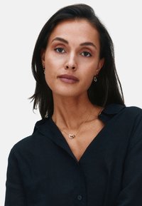 Moteris su tamsiais plaukais, dėvinti tamsią marškinių tipo palaidinę, auksinę širdies formos vėrinį ir atitinkančius auksinius auskarus, žiūri tiesiai į priekį.