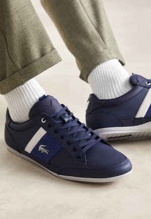 Sneakers casual blu navy con striscia bianca e logo, indossate con calzini bianchi a coste e pantaloni beige risvoltati su pavimento chiaro.