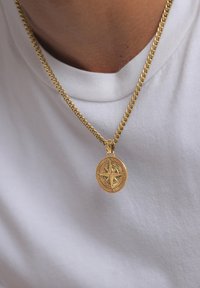 Collana a catena dorata con un pendente a forma di bussola rotonda, caratterizzata da indicatori direzionali e una superficie testurizzata, indossata sopra una camicia bianca.