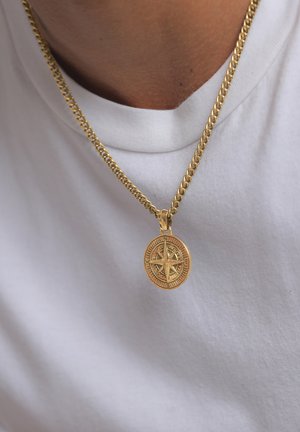 Collana a catena dorata con un pendente a forma di bussola rotonda, caratterizzata da indicatori direzionali e una superficie testurizzata, indossata sopra una camicia bianca.