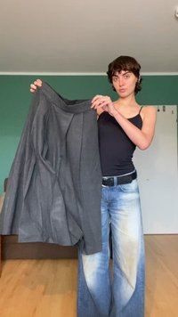 Szara plisowana spódnica z lekkiego materiału, z wysokim stanem, przedstawiona na tle niebieskich dżinsów i gładkiej ściany.