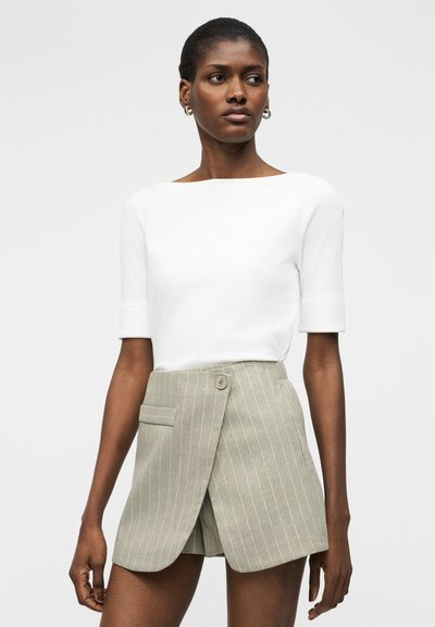 Γυναίκα με λευκό τοπ με κοντά μανίκια και μπεζ σορτς με ρίγες, εμπρός τύπου wrap, με κουμπί και λεπτομέρειες τσέπης.