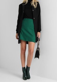 Manteau noir sur un col roulé noir, mini-jupe verte et bottines vernies vertes. La tenue comprend un sac noir avec une chaîne dorée.