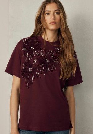 Jeune femme aux longs cheveux portant un t-shirt bordeaux décoré de motifs floraux texturés et de petites perles sur la poitrine.