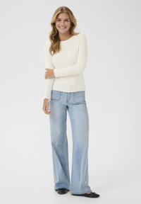 Crème ribbelige langwerpige top in combinatie met lichtblauwe wijde jeans. De top heeft een subtiele scalloped zoom en een aansluitende pasvorm. Zwarte ballerina's.
