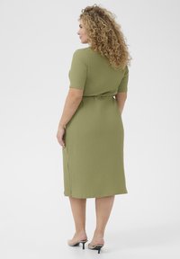Olivegrünes Kleid mit strukturiertem Finish, kurzen Ärmeln, tailliertem Schnitt und einem Seitenschlitz, kombiniert mit nudefarbenen High Heels.