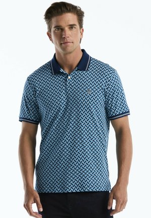 Man wearing blue patterned polo shirt with dark collar and sleeve trim standing against white background.