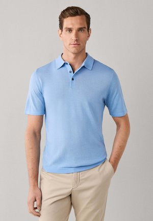 Man with short brown hair wearing a light blue short-sleeve polo shirt and beige pants, standing with one hand in pocket against plain background.